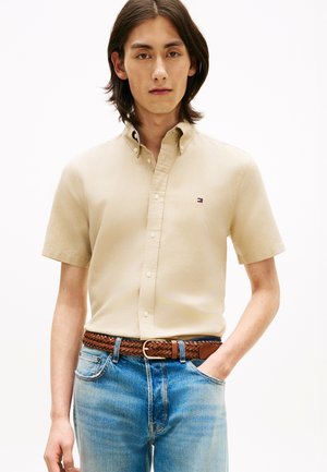 Young man with shoulder-length dark hair wearing a beige short-sleeve button-up shirt, braided brown leather belt, and blue jeans.