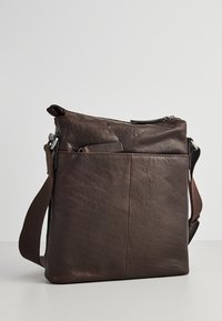 Mørkebrun læder crossbody taske med frontlomme og justerbar strop, stående oprejst mod en ensfarvet baggrund.