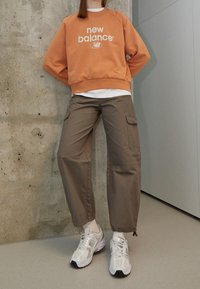 Felpa arancione con logo bianco, abbinata a pantaloni cargo beige con tasche laterali. Sneakers grigie con accenti bianchi.