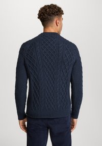 Mężczyzna z kręconymi włosami, ubrany w ciemnoniebieski sweter w sploty oraz granatowe spodnie, stojący tyłem do kamery na jednolitym tle.