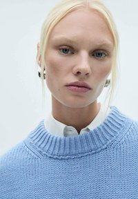 Hellblauer Strickpullover mit einem gerippten Kragen, über einem weißen Hemd mit Kragen getragen, das die strukturierten Muster und das entspannte Design hervorhebt.