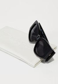 Sorte oversized solbriller med en glatt finish og mørke linser, plassert på et hvitt textured brilleetui med preget merkevare.
