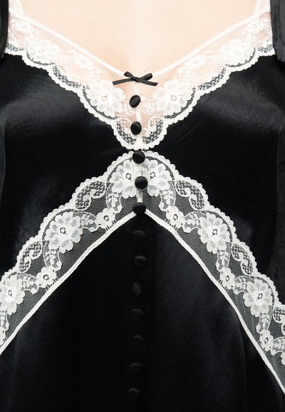 Μαύρο βελούδινο ένδυμα με λευκή δαντέλα με σχέδιο λουλουδιών και μικρά μαύρα κουμπιά κατά μήκος του μπροστινού μέρους, με μικρό μαύρο φιόγκο στο λαιμό.