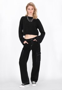 Schwarzer verkürzter Pullover mit langen Ärmeln, kombiniert mit schwarzen Cargo-Hosen mit Seitentaschen und weißer Naht, getragen mit schwarzen Sneakern.