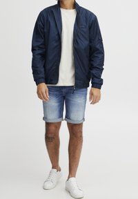 Marineblauwe lichtgewicht jas met een rits aan de voorkant, elastische manchetten, gedragen over een licht T-shirt en denim shorts. Witte sneakers maken de look compleet.