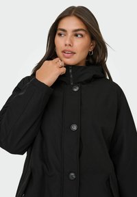 Juodas su gobtuvu striukė su užtrauktuku ir dideliais mygtukais. Yra lygi išorinio audinio danga ir šoninės kišenės, skirtos komfortui ir šilumai.