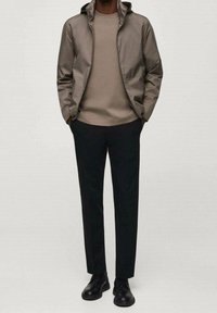 Leichte, braune Jacke mit Kapuze, kombiniert mit einem beigen Pullover und schwarzen Hosen. Schwarze Schuhe vervollständigen das Outfit und verleihen einen eleganten Look.