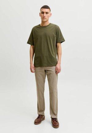 Mlad moški stoji, oblečen v olivno zeleno majico, bež hlače in rjave usnjene čevlje na enostavni svetli podlagi.