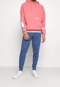 Sudadera rosa con logo blanco y mangas a rayas multicolor, combinada con pantalones de chándal azules con franjas laterales blancas y zapatillas blancas.
