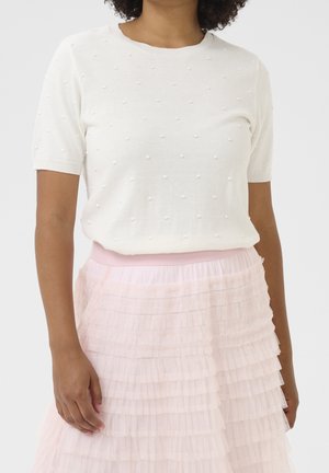 Kobieta ubrana w biały top z krótkim rękawem z małymi teksturowanymi kropkami, zestawiony z jasnoróżową, warstwową tiulową spódnicą.