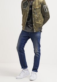 Man som bär en olivgrön bomberjacka med lappar, mörkgrå t-shirt, blå jeans och vita sneakers som står mot en enfärgad bakgrund.