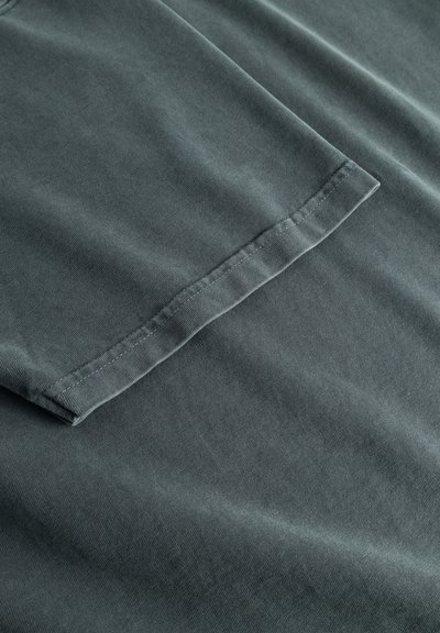 T-shirt in tessuto grigio scuro che mostra una texture liscia, cuciture piatte sulle maniche e una leggera lucentezza dovuta alla qualità del materiale.