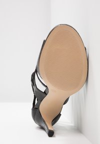 Sandale noire à talon haut avec une semelle texturée et un talon fin, montrée par dessous sur fond blanc et coin de mur.
