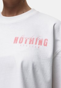 Felpa bianca in cotone, caratterizzata da una grafica testurizzata e usurata in rosa con la scritta "NOTHING" e "atelier" in un font moderno.
