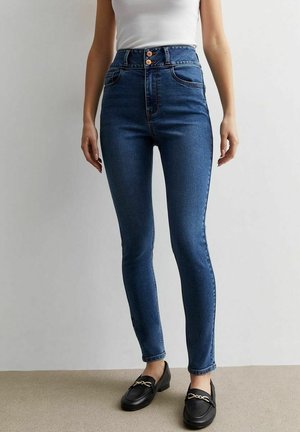 Smaltsiddende blå jeans med høj talje og dobbelt knaplukning, forlommer, båret med sorte loafers og en hvid ærmeløs top.