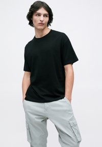 Seorang pemuda mengenakan kaos hitam lengan pendek dan celana kargo abu-abu terang dengan tangan dimasukkan ke saku, memandang ke sisi di depan latar belakang polos.