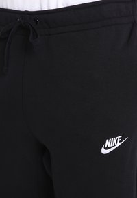 Pantaloni della tuta neri con cintura elastica e lacci. Presentano il logo Nike bianco sul lato sinistro. Tessuto morbido con una finitura liscia.