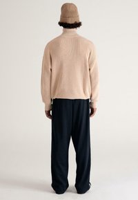 Mann mit beiger Strickmütze und passendem gerippten Rollkragensweater sowie lockeren schwarzen Hosen, der sich mit dem Rücken zu einer schlichten Hintergrundwand steht.