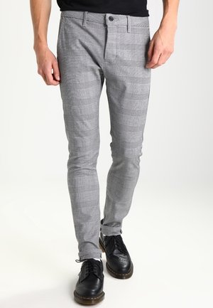 Hombre con pantalones ajustados de cuadros grises y zapatos brogue de cuero negro, de pie frente a un fondo liso.