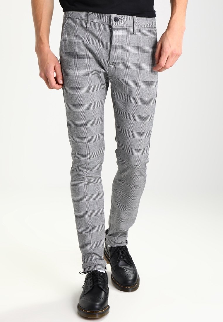 Homme portant un pantalon slim à carreaux gris et des chaussures brogues en cuir noir, debout devant un fond uni.
