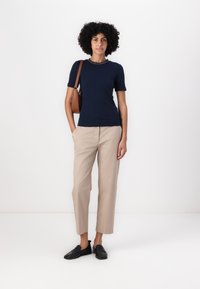 Top scurt cu maneci, de culoare bleumarin, cu un guler în contrast, asortat cu pantaloni bej deschis, croiți, și mocasini negri, ținând o geantă de umăr maro.