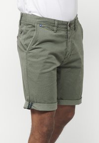 Vīriešu olīvu zaļie teksturētie kokvilnas šorti ar sarullētām atloka malām, valkāti ar baltu krekli, redzami līdz vidus cīpslim un gurniem.