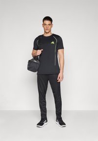 Sort atletisk t-shirt med grønne accenter og hvid piping, parret med sorte bukser og sorte sneakers, mens der bliver holdt en lille sort taske.