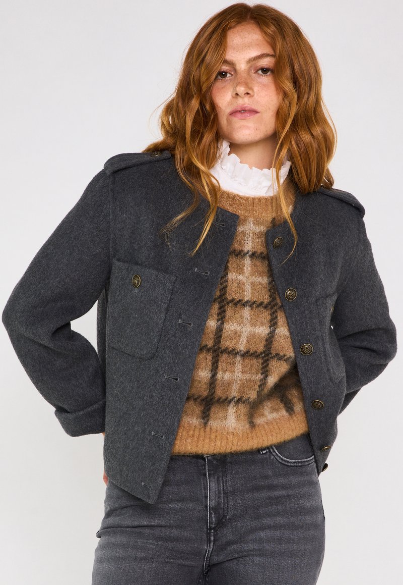 Veste en laine grise avec fermeture à bouton et épaulettes, dotée d'une poche avant et portée par-dessus un pull écossais beige.