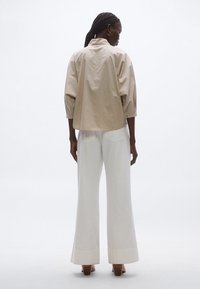 Cămașă supradimensionată cu dungi bej, mâneci trei sferturi, vedere din spate. Combinate cu pantaloni albi largi, talie înaltă și pantofi maro cu toc gros.