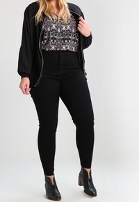 Schwarze Bomberjacke über einer gemusterten Bluse, kombiniert mit hoch taillierten schwarzen Hosen und Stiefeletten mit Ausschnitt und Holzabsatz.
