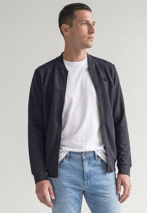 Homme portant une veste noire zippée sur un t-shirt blanc et un jean bleu clair, regardant vers la droite sur un fond uni.