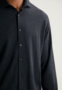 Hombre vistiendo una camisa de manga larga abotonada gris oscuro con un sutil logo bordado en el lado izquierdo del pecho, contra un fondo liso.