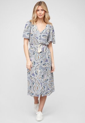 Blondínka v modro-béžových midi šatách s paisley vzorom, krátkymi rukávmi a bielymi teniskami stojaca pred jednofarebným sivým pozadím.