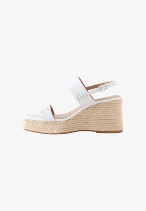 Pies con sandalias blancas de plataforma con suelas tejidas, combinadas con un dobladillo de falda a cuadros azul y blanco visible por encima de los tobillos.