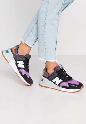 Person wearing light blue cropped jeans and black, purple, and white New Balance sneakers with gum soles standing on a white floor.