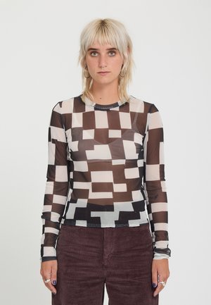 Jeune personne aux cheveux blond clair porte un haut transparent à motif géométrique noir et blanc et un pantalon en velours côtelé marron foncé.