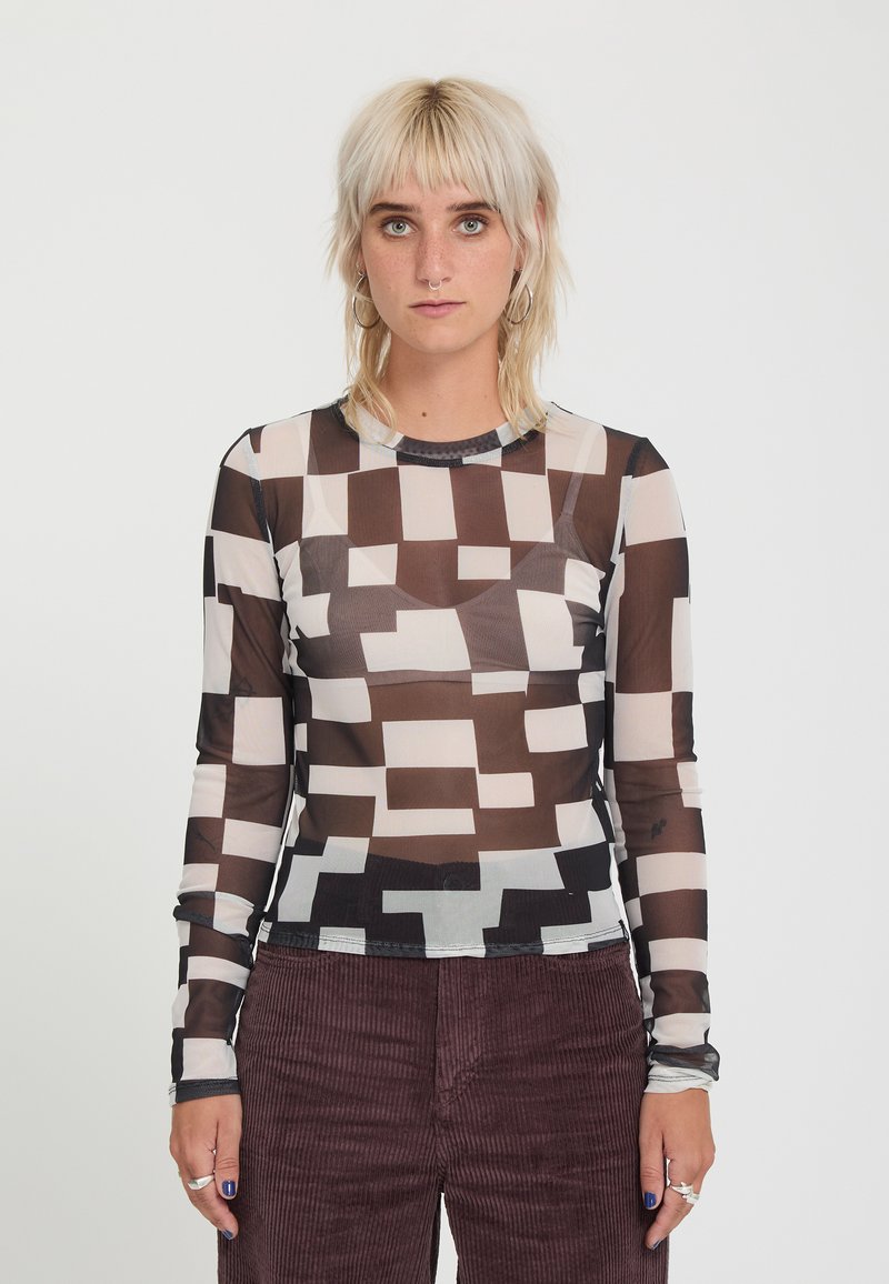 Jeune personne aux cheveux blond clair porte un haut transparent à motif géométrique noir et blanc et un pantalon en velours côtelé marron foncé.