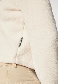 Fleece-takki vaalean kremän värissä, jossa on teksturoitu pinta ja musta sivutagi. Yhdistetty beiget housut.