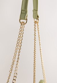 Gros plan sur des sangles en chaîne dorée attachées à des boucles en cuir vert clair sur un sac, sur un fond neutre.