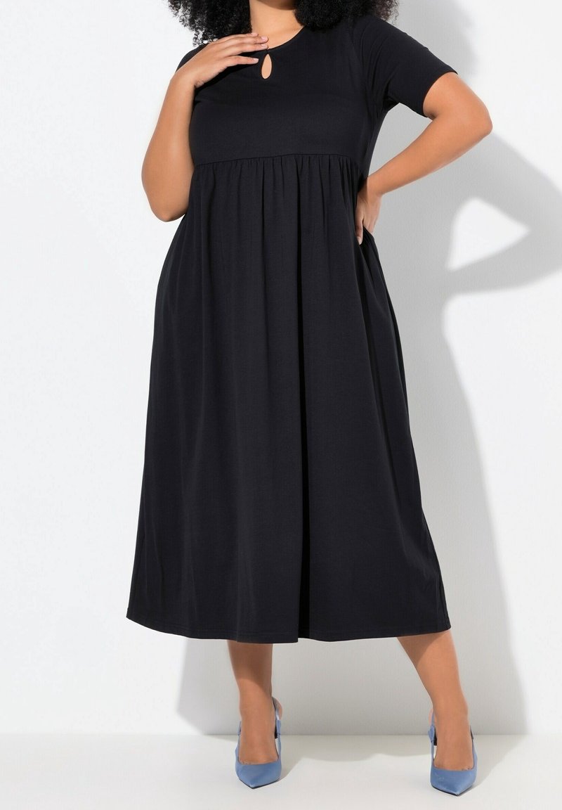 Femme portant une robe midi noire à manches courtes avec une petite ouverture devant et des talons bleus à bout pointu, posant avec la main sur la poitrine et la taille.