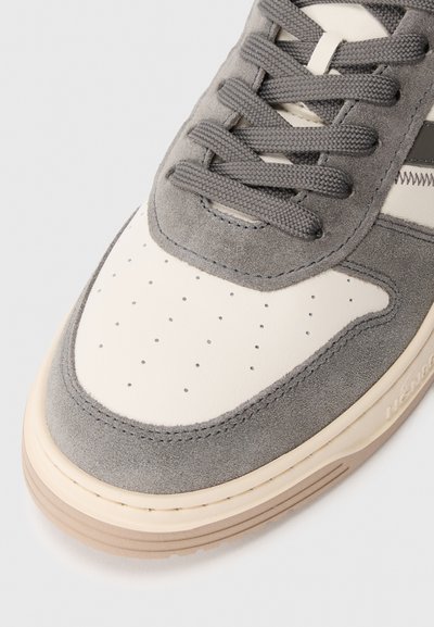 Sneaker z szaro zamszowym wierzchem, kremową skórzaną czubkiem i perforowanymi detalami. Posiada szare sznurowadła oraz grubą beżową gumową podeszwę.