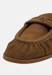 Scarpa slip-on in suede marrone con texture arricciata, punta rotonda e un accento dorato sulla parte superiore. Suola piatta di colore abbinato.