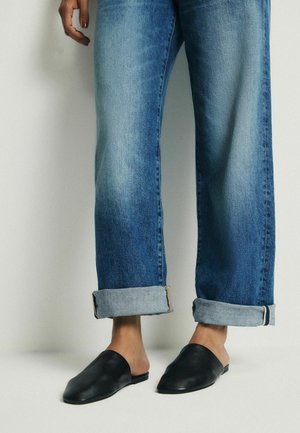 Person iført manchetterede blå jeans og sorte slip-on flade sko, stående mod en ensfarvet lys baggrund.