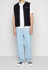 Gilet in pile nero con zipper frontale, dotato di tasche laterali e una tasca sul petto. Indossato sopra una t-shirt bianca e jeans larghi di colore azzurro chiaro.