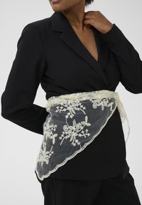 Spetsband i krämfärg med blommönster, bärs över en svart kavaj. Har en vågig kant och mjuk textur.