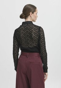 Camicia di pizzo nero con colletto, maniche lunghe e un motivo testurizzato, abbinata a pantaloni bordeaux a righe.