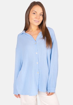 Junge Frau mit langen braunen Haaren, die ein lockeres hellblaues Hemd und weiße Hosen trägt, steht vor einem schlichten Hintergrund.