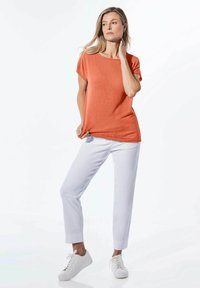 Oranje T-shirt met korte mouwen en een ronde halslijn, gecombineerd met witte rechte broek en witte sneakers op een effen achtergrond.