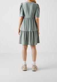 Hellgrünes, kurzärmliges Kleid mit einem gestuften Design, das ein Zickzackmuster und subtile Texturdetails aufweist. Kombiniert mit beigen Schuhen und weißen Socken.