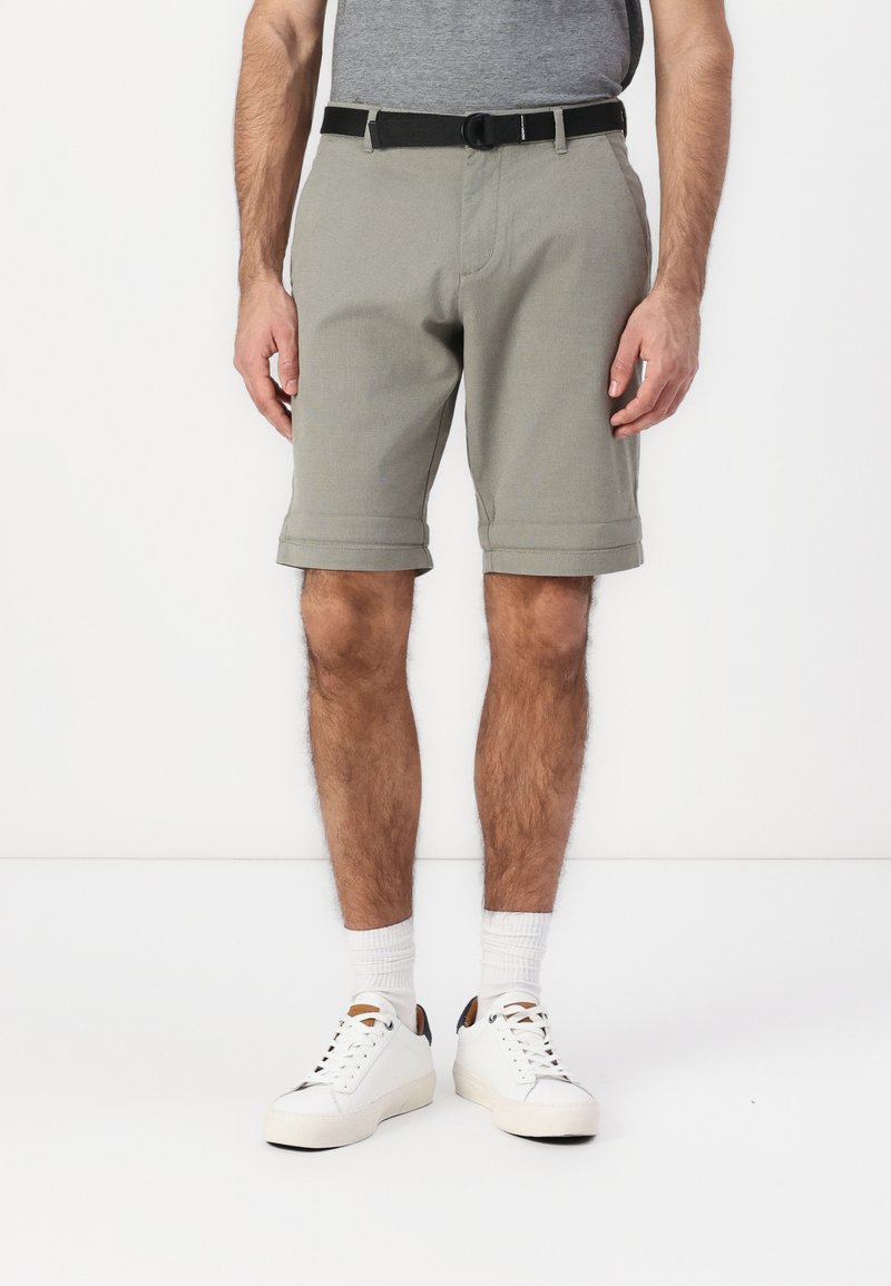 Shorts gris en tissu avec des ourlets retournés, dotés d'une ceinture noire. Portés avec des baskets blanches et des chaussettes blanches côtelées.